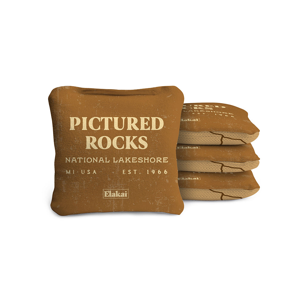 #color_tan A set of four beige travel-size cornhole bags with 'PICTURED ROCKS NATIONAL LAKESHORE MI USA EST. 1966' printed on them, placed against a white background.