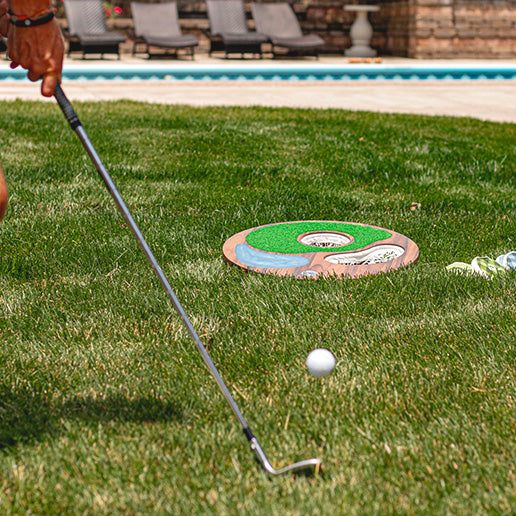 Person playing island hole mini golf on a grassy area with a pool in the background