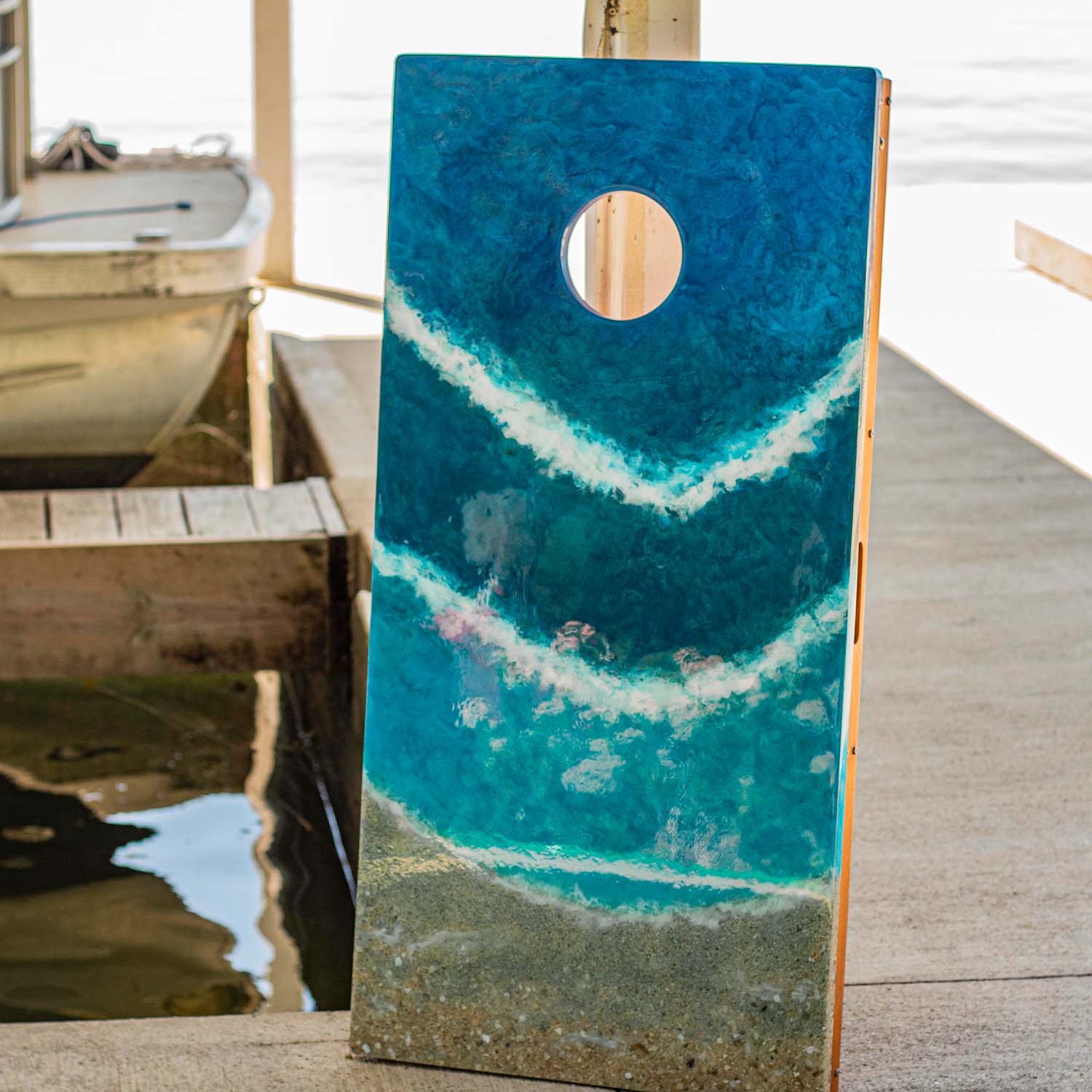 Beach resin cornhole board displayed on a lakeside dock with boat in background