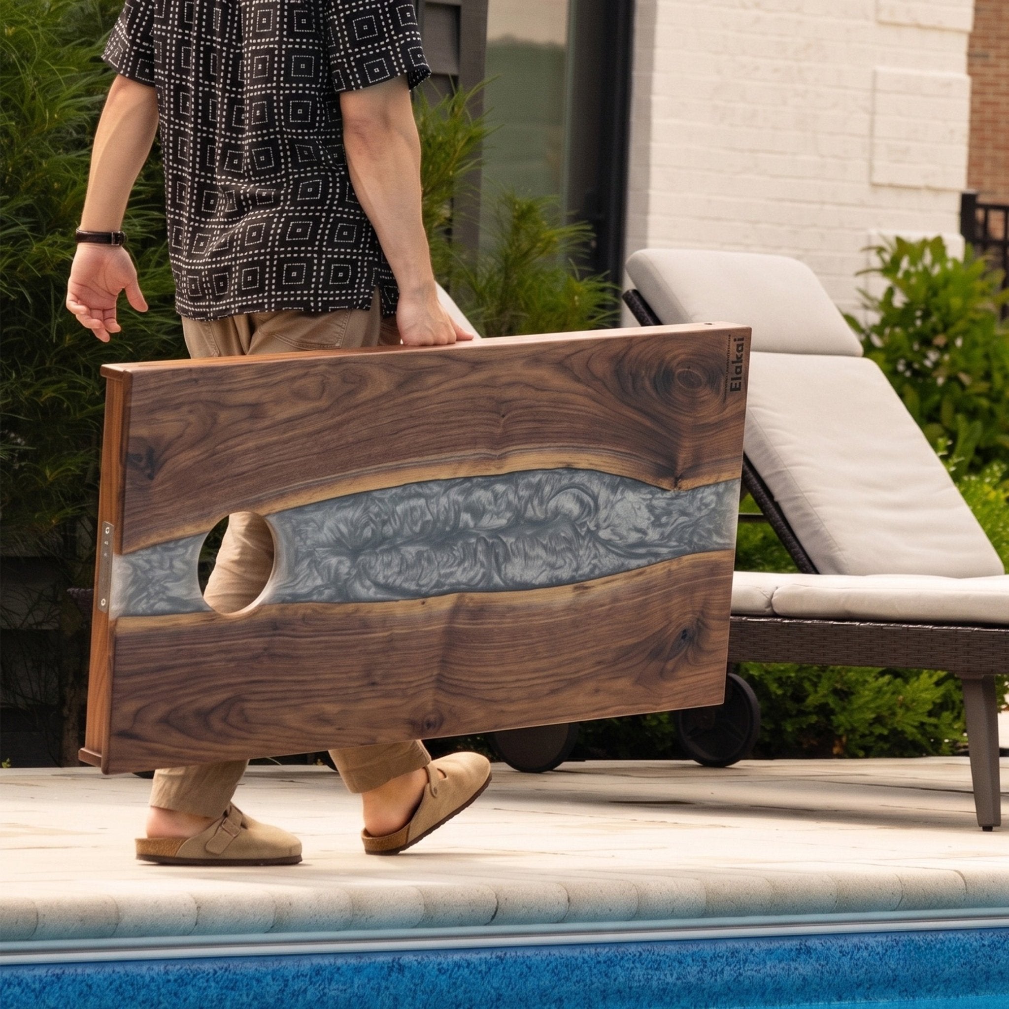A person carries a wooden cornhole board with a wavy gray resin inlay and a circular cutout, photographed outdoors beside a pool with a lounge chair in the background. #color_gray stone
