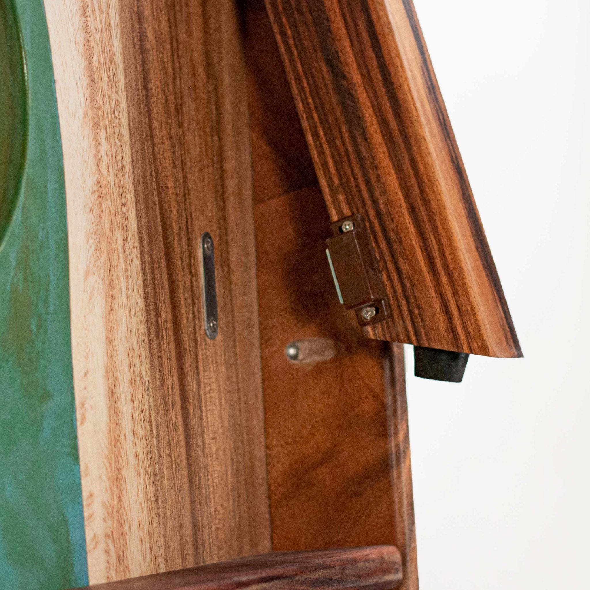 Close-up detail of a wooden cornhole board, showing rich wood grain, a metal latch, and a small black rubber stopper against a white background. #color_teal and orange