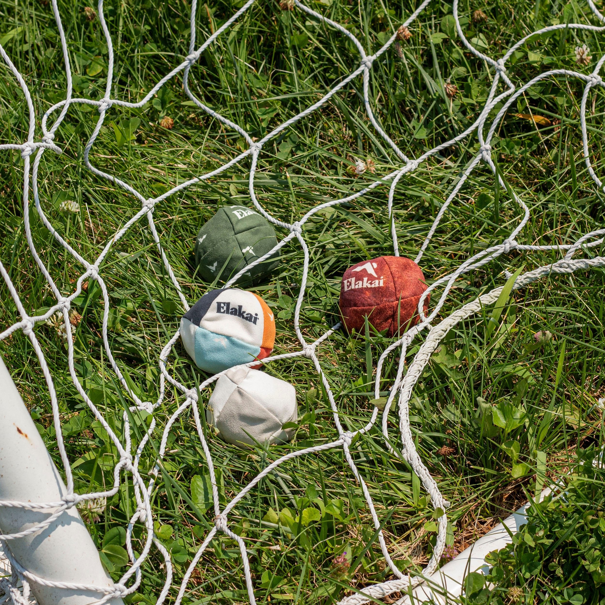 Four Elakai brand balls hanging on a soccer net against a grassy background
