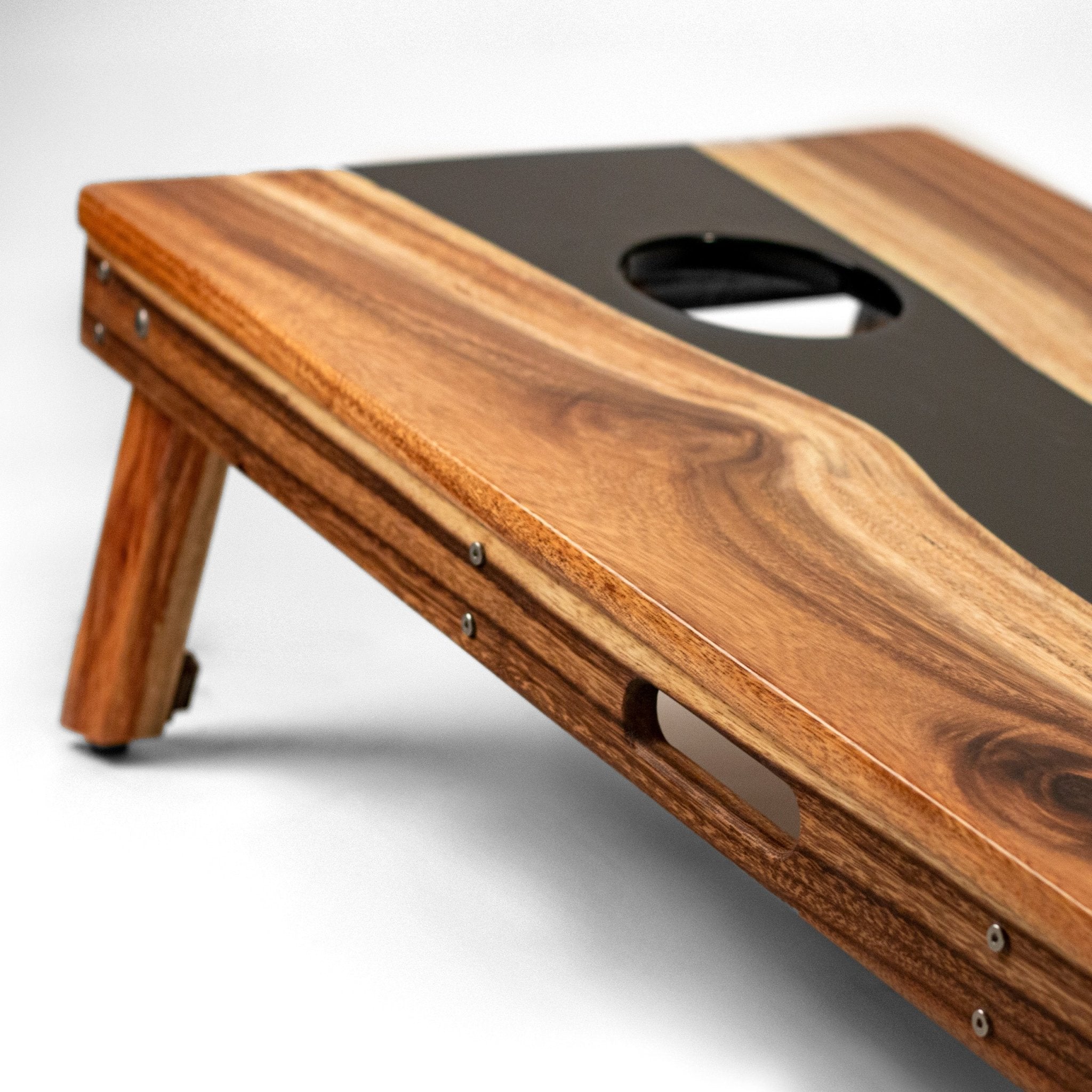 A close-up of a wooden cornhole board with a live-edge wood grain finish and a dark center strip, shown at an angle on a white background. #color_black