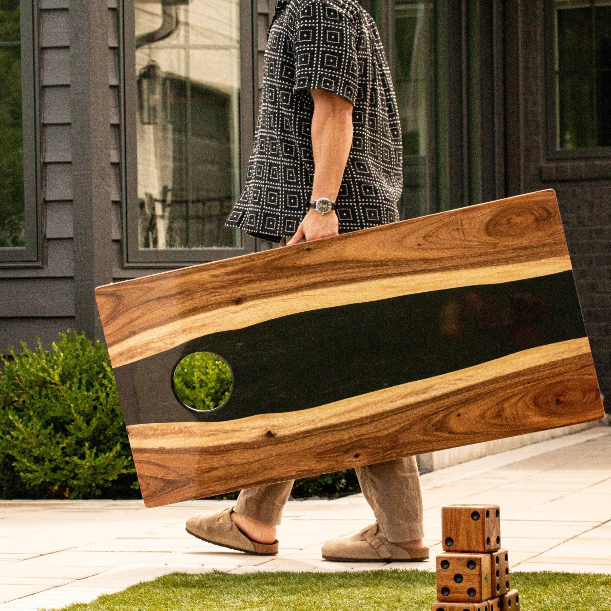 A person outdoors carrying a large rectangular cornhole board with a black center stripe and a circular cutout, with stacked wooden dice on grass nearby and a house in the background. #color_black