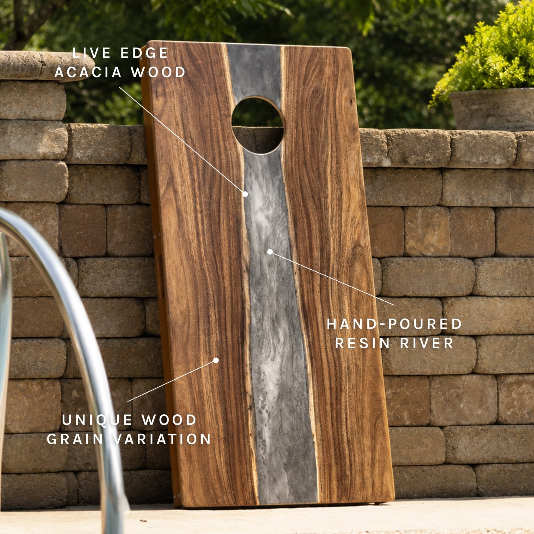 A large rectangular wooden cornhole board with a circular cutout, leaning against a stone wall outdoors. White text callouts point to features including live edge acacia wood, hand-poured resin river, unique wood grain variation, and natural live edge. #color_gray stone