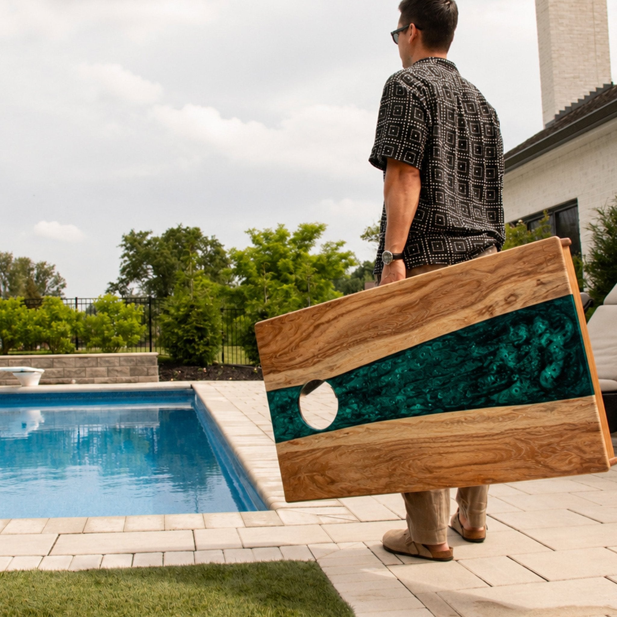 A man carrying a large wooden board with a green marbled inlay and a circular hole, standing beside a swimming pool in a backyard. #color_green pearl