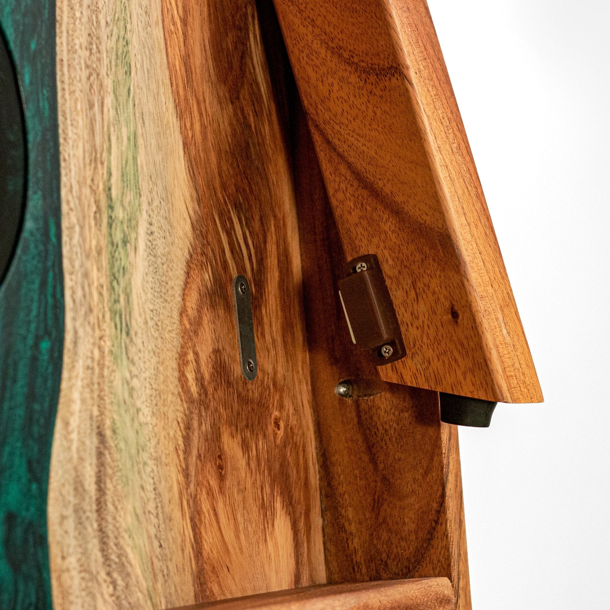 Close-up of a wooden cornhole board, showing warm medium-brown wood grain, a dark metal hinge, and part of a green-and-brown patterned surface on the left against a white background. #color_green pearl