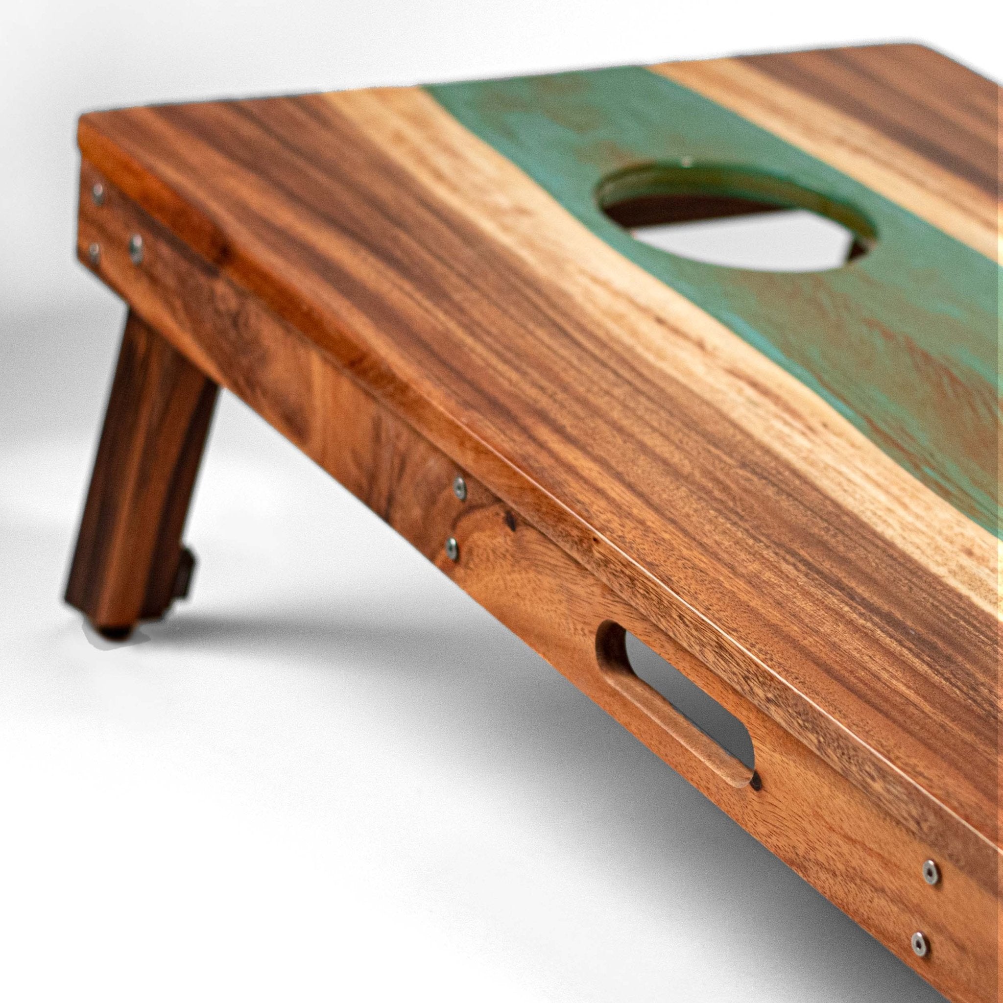 A close-up of a wooden cornhole board with a green resin inlay, metal corner brackets, and a cutout handle, photographed on a light gray studio background. #color_teal and orange