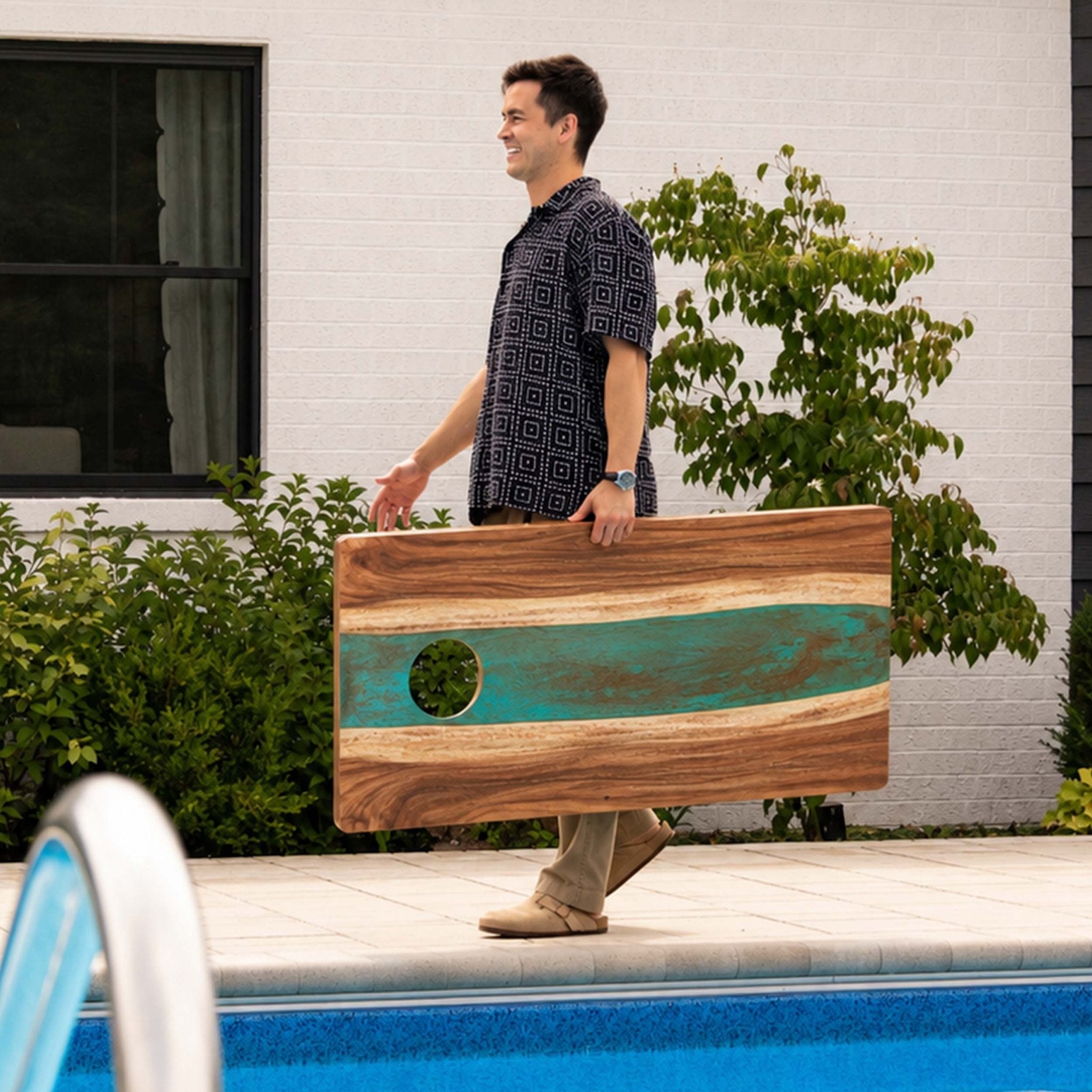 A man walks beside a swimming pool carrying a large wooden cornhole board with a turquoise inlay and a circular cutout. The setting is outdoors in front of a white brick wall and green shrubs. #color_teal and orange